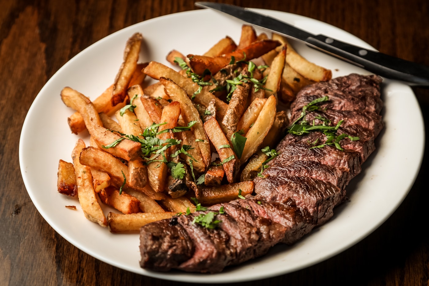 Steak frites du jour : assiette blanche, frites maison à la persillade, couteau posé sur la table en bois sombre.