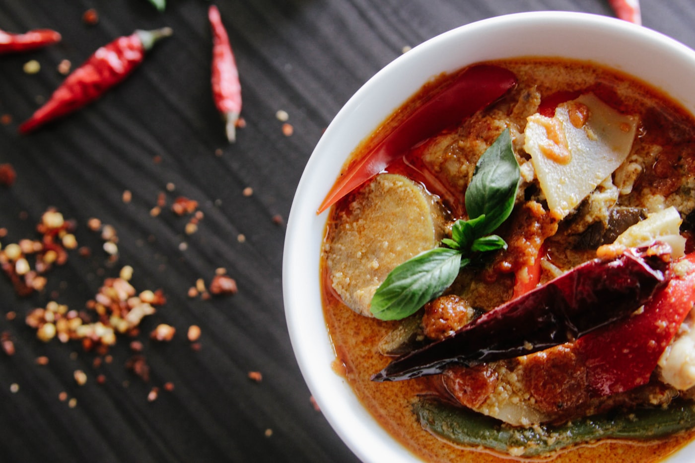Curry rouge vu du dessus dans un bol blanc posé sur fond noir, piments séchés et épices éparpillés tout autour.