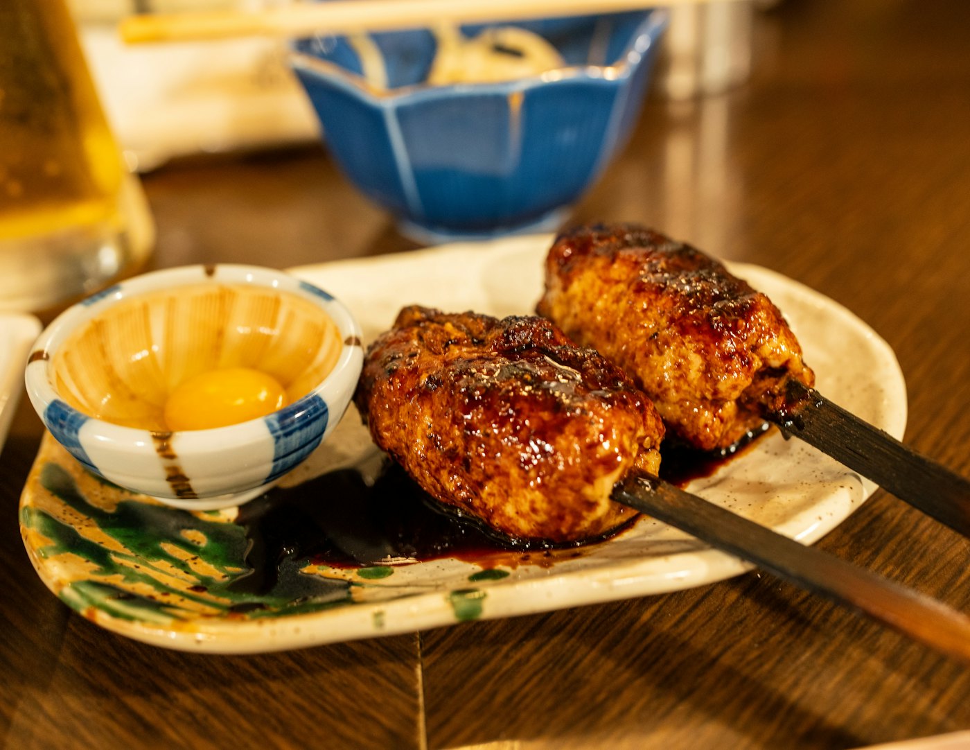 Deux brochettes de tsukune laquées sur une petite assiette, accompagnées d'un bol de jaune d'œuf, posées sur le comptoir