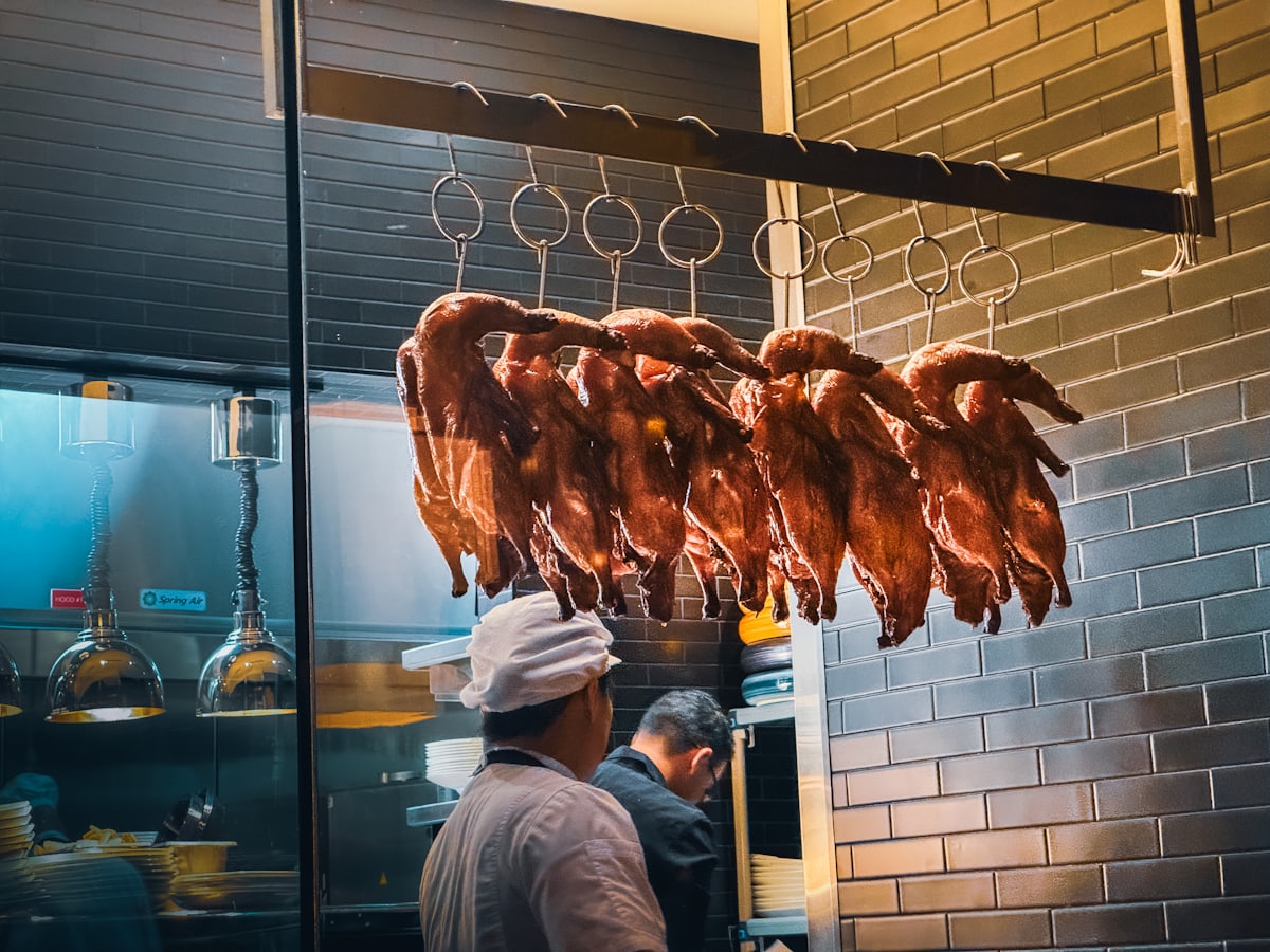 Dans la cuisine, une rangée de canards laqués suspendus à leurs crochets devant le mur carrelé gris-bleu, cuisiniers au travail derrière.
